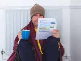 Person holding a winter electric bill and hot coffee, wrapped in a blanket beside a radiator, worried about high heating costs